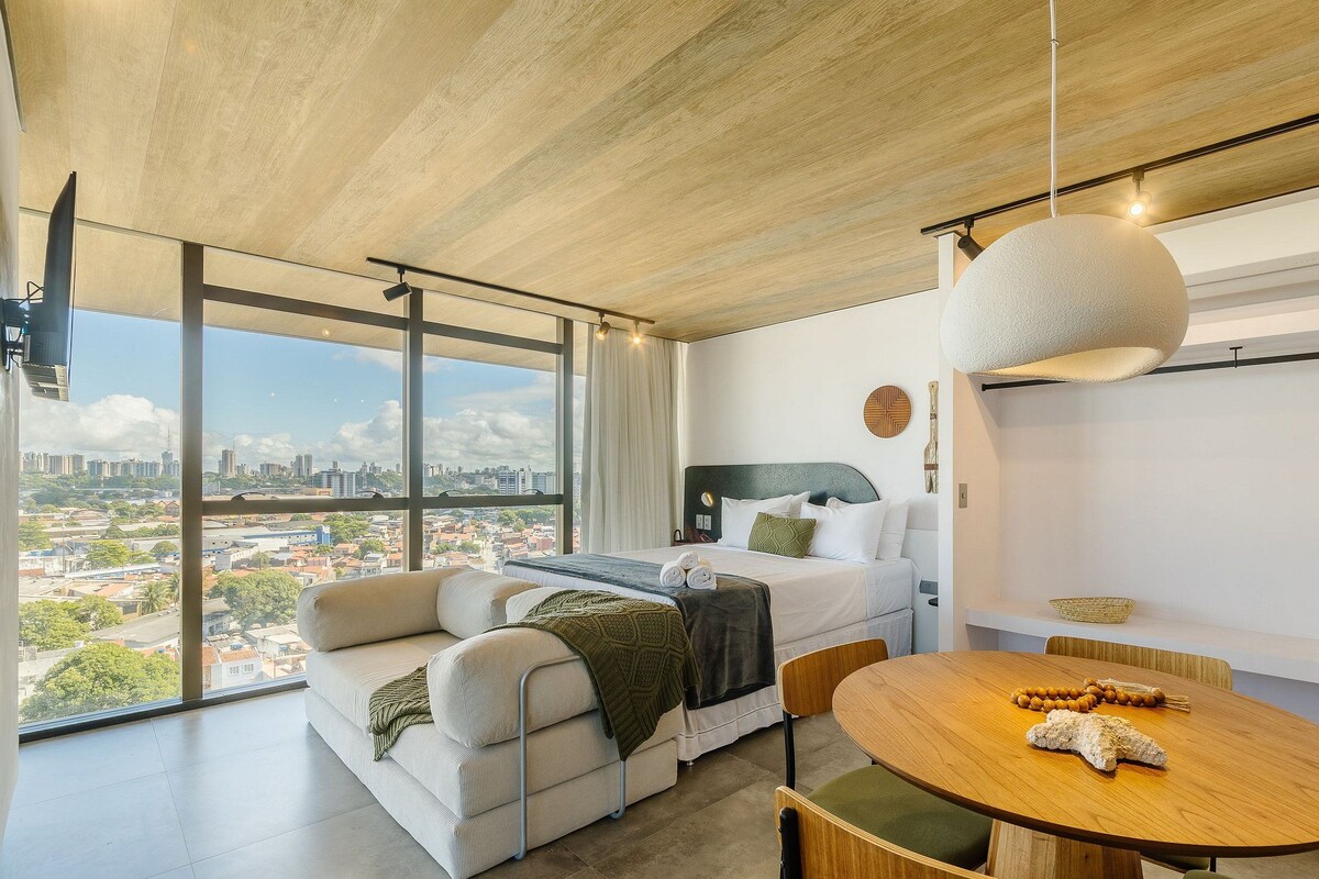 Una habitación de hotel moderna con un ventanal grande, un sofá, una mesa de comedor redonda y una lámpara colgante.