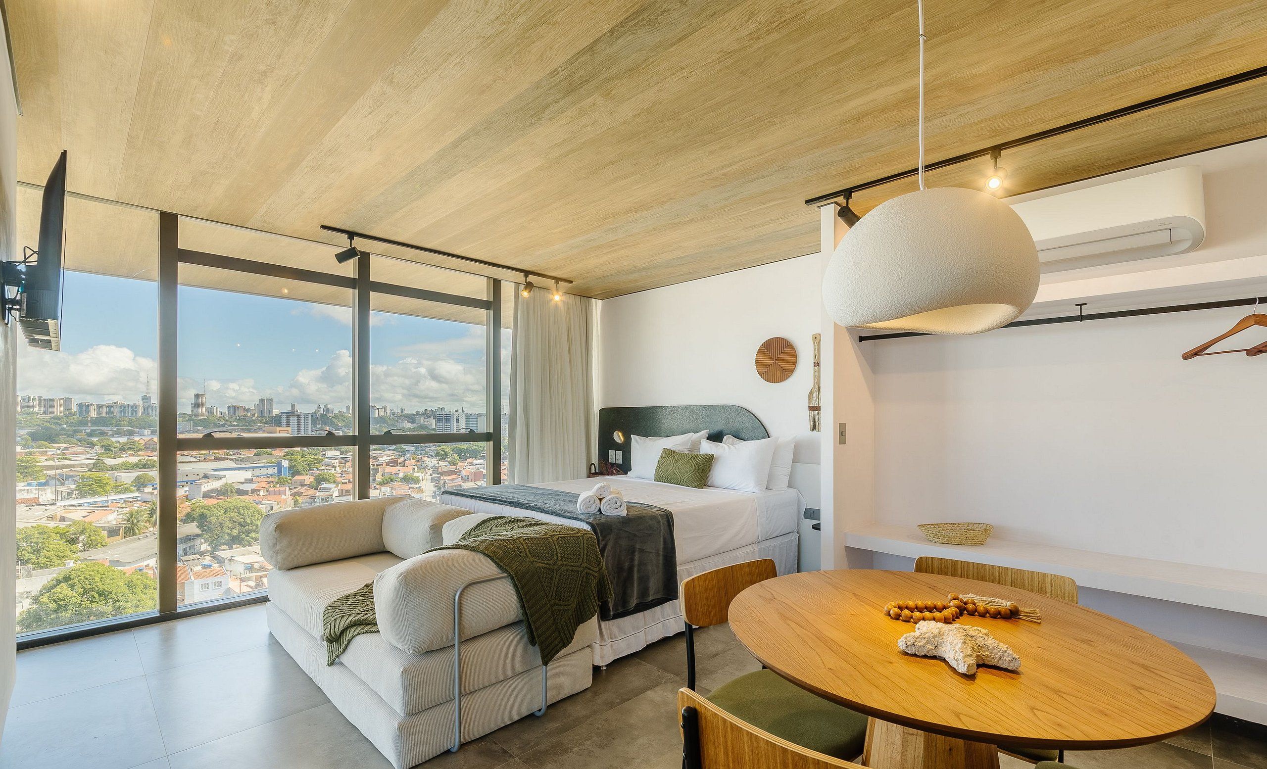 A modern hotel room with a large window, a sofa, a round dining table, and a hanging light fixture.
