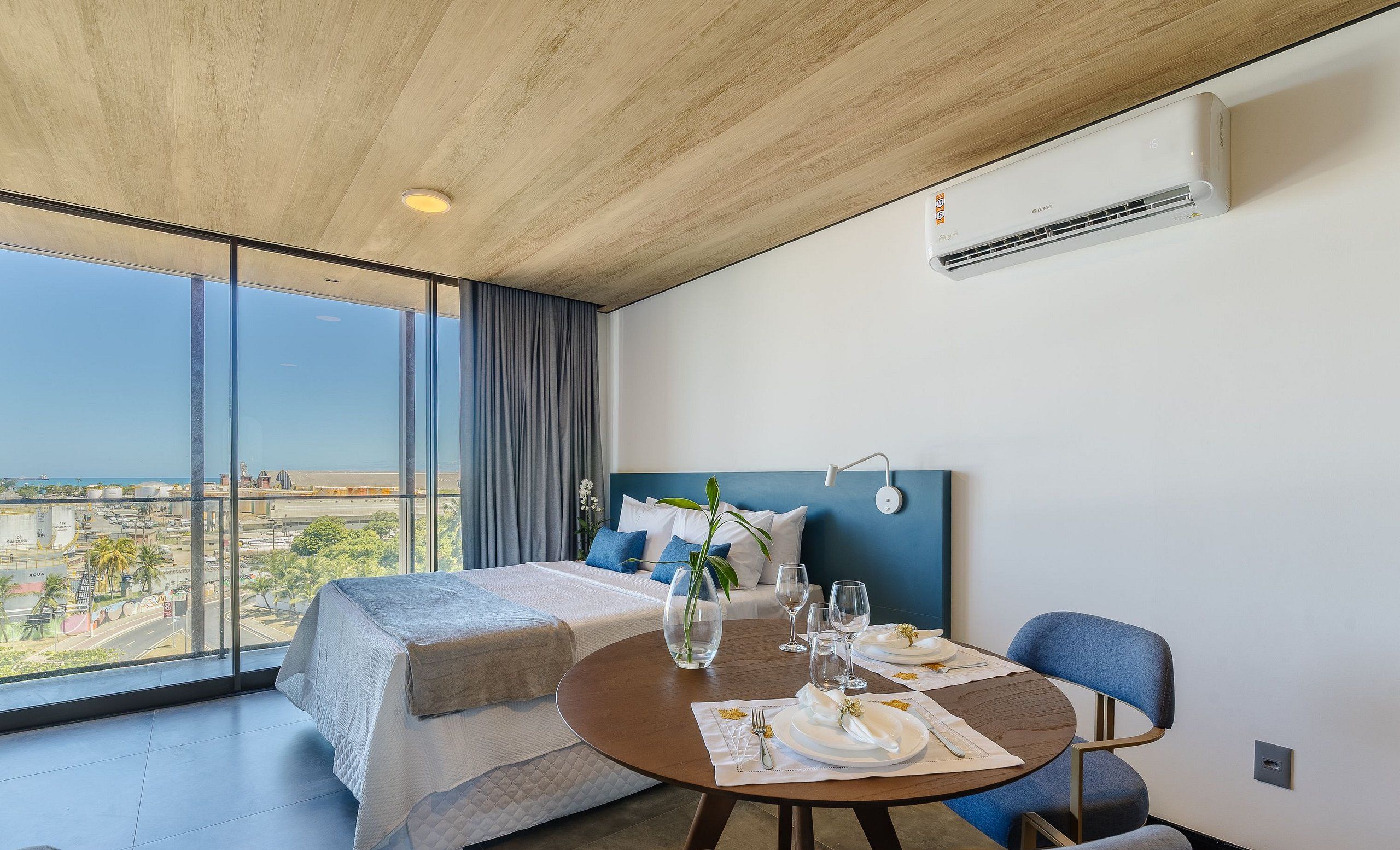 A hotel room with a large window, a bed, a dining table set for two, and an air conditioning unit.