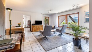 Una sala de estar moderna con televisor, asientos, zona de comedor y vistas amplias a través de grandes ventanales.