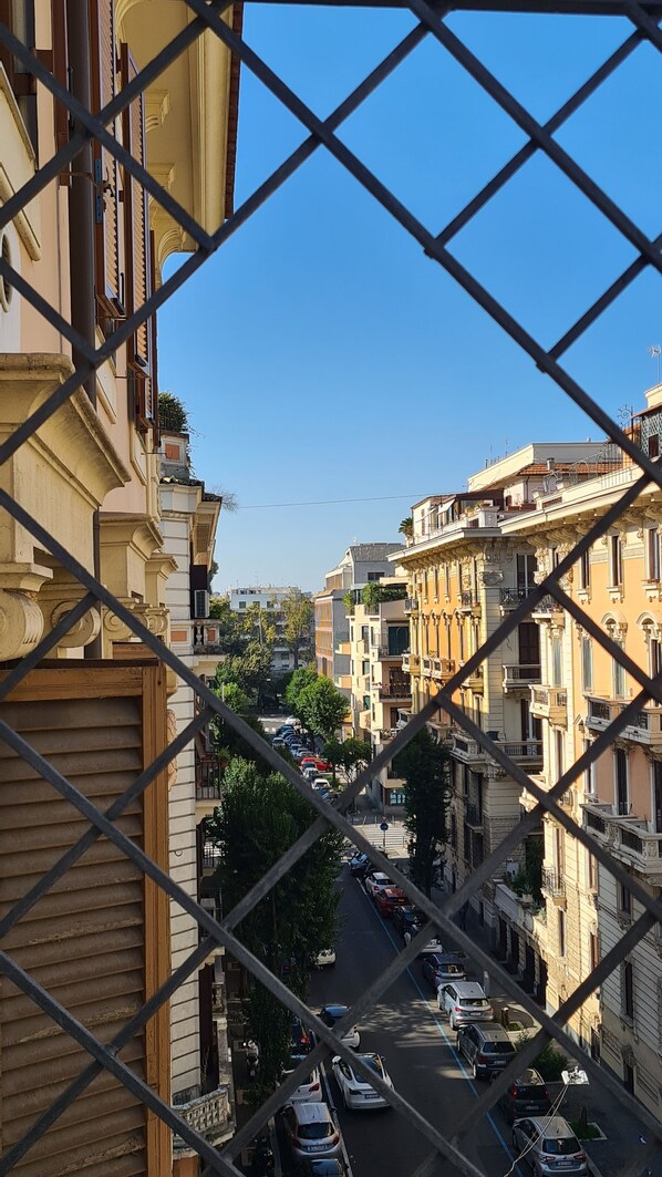 A street view through a metal fence with buildings on either side, cars parked along the street, and a clear blue sky.