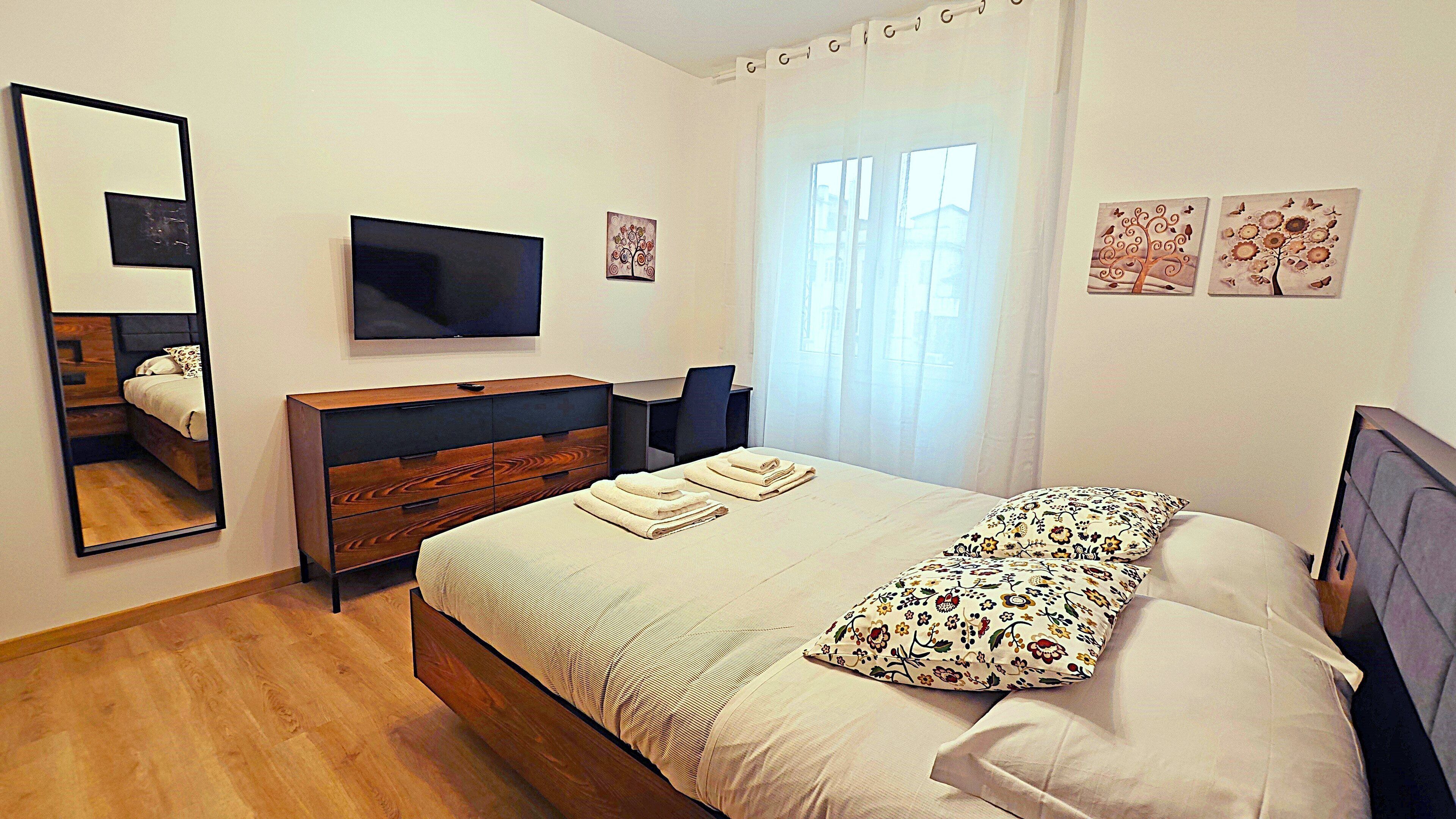 Un dormitorio moderno con una cama de madera, un televisor de pantalla plana, un escritorio y un amplio ventanal con cortinas translúcidas.