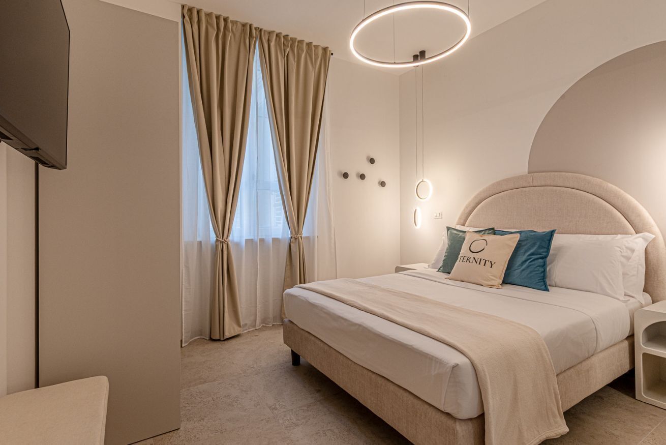 A modern bedroom with a large bed, beige curtains, a round light fixture, and a wall-mounted TV.