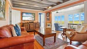A spacious living room with a wooden ceiling, a large flat-screen TV, a fireplace, and a view of the outdoors. - Historic Marshfront St Simons Estate • Heated Pool • Private Dock (St Simons)