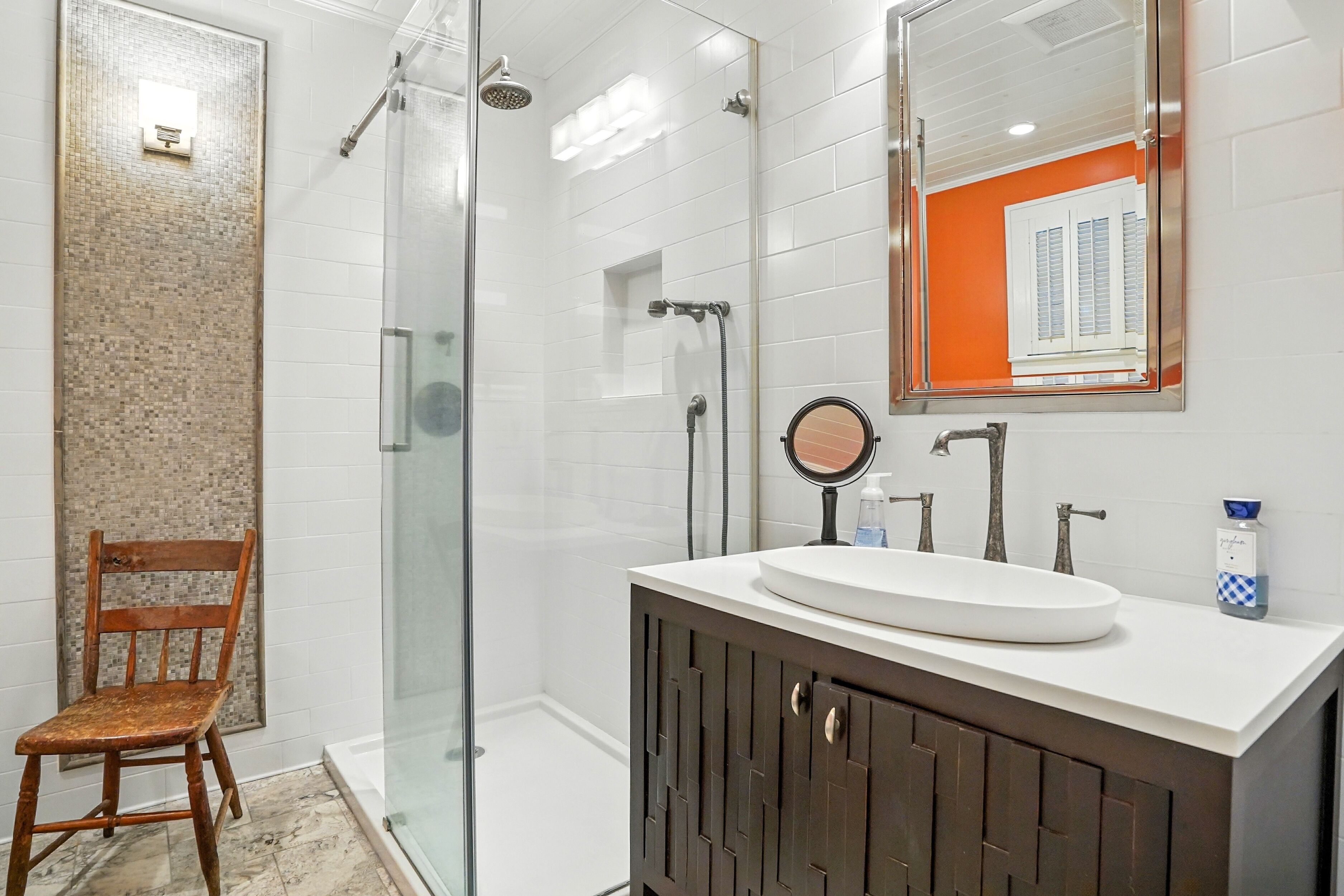 A modern bathroom with a glass-enclosed shower, a wooden chair, a white sink with a round mirror, and a dark wooden cabinet.