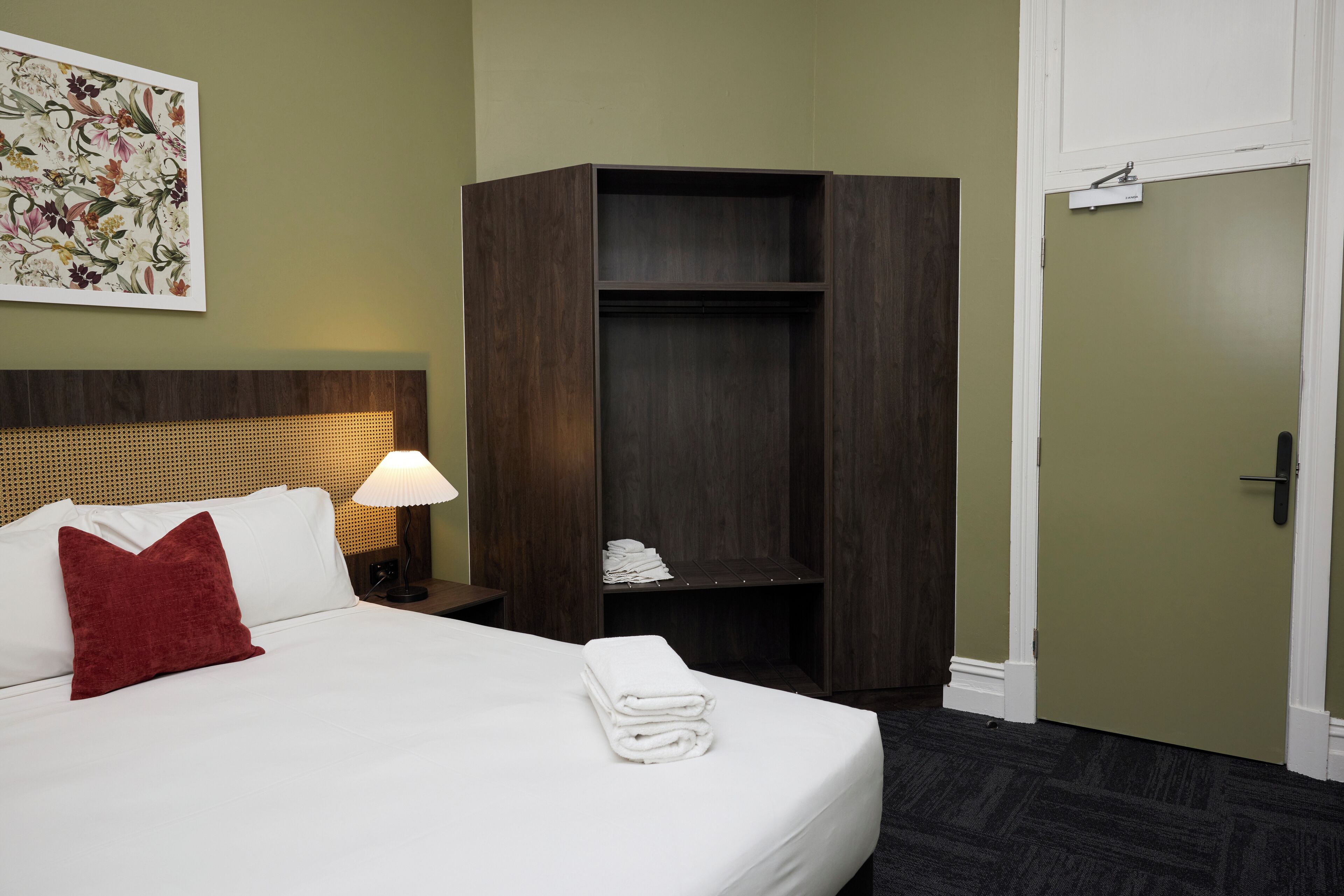 A neatly made bed with white linens and a red pillow, a wooden wardrobe, a wall-mounted lamp, and a door with a nameplate.