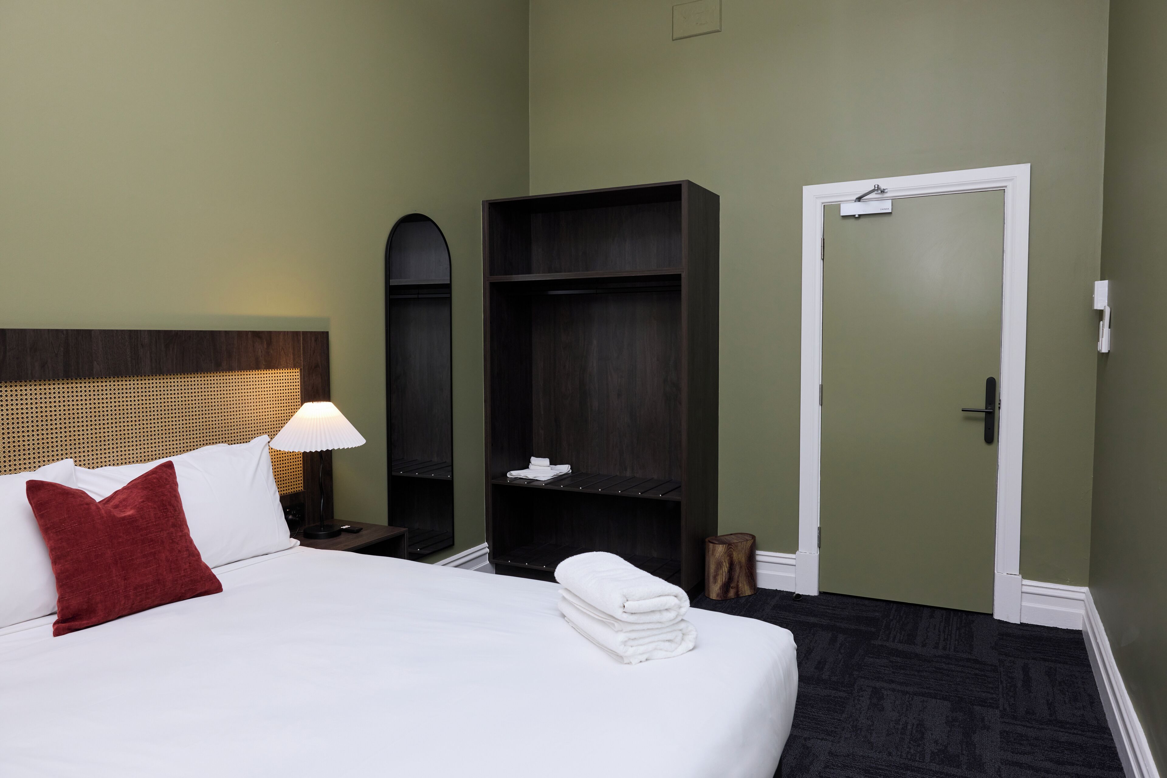 A hotel room with a bed, a red pillow, a lamp, a wardrobe, and a door.