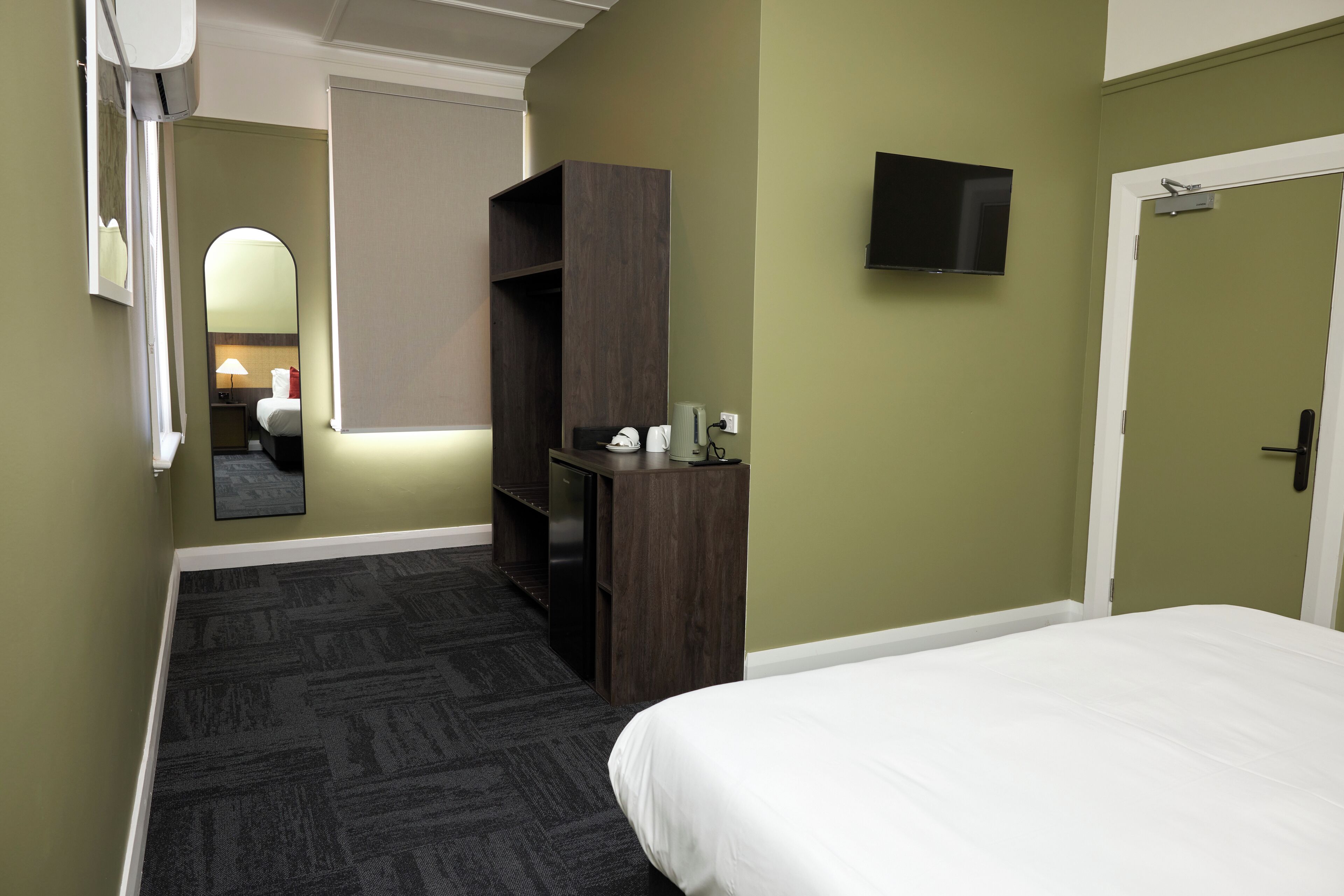 A hotel room with a bed, a desk, a mirror, a TV, and a door.