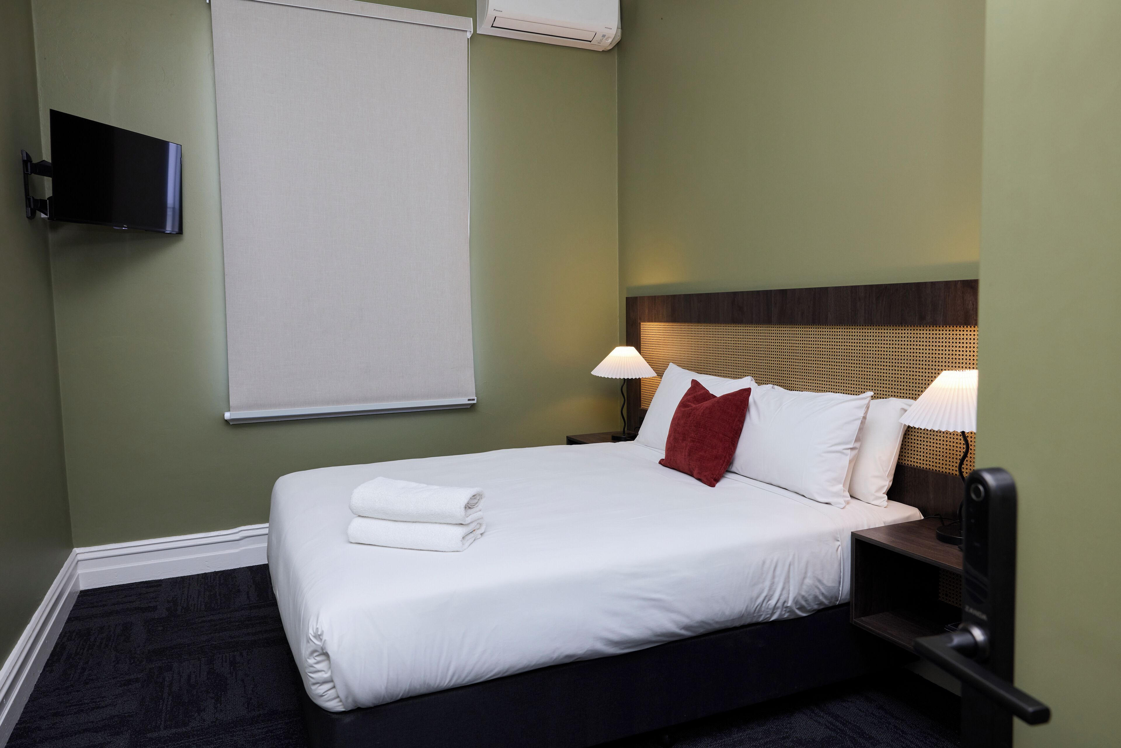 Una cama bien hecha con sábanas blancas y una almohada roja, un televisor de pantalla plana montado en la pared y una ventana con persiana.