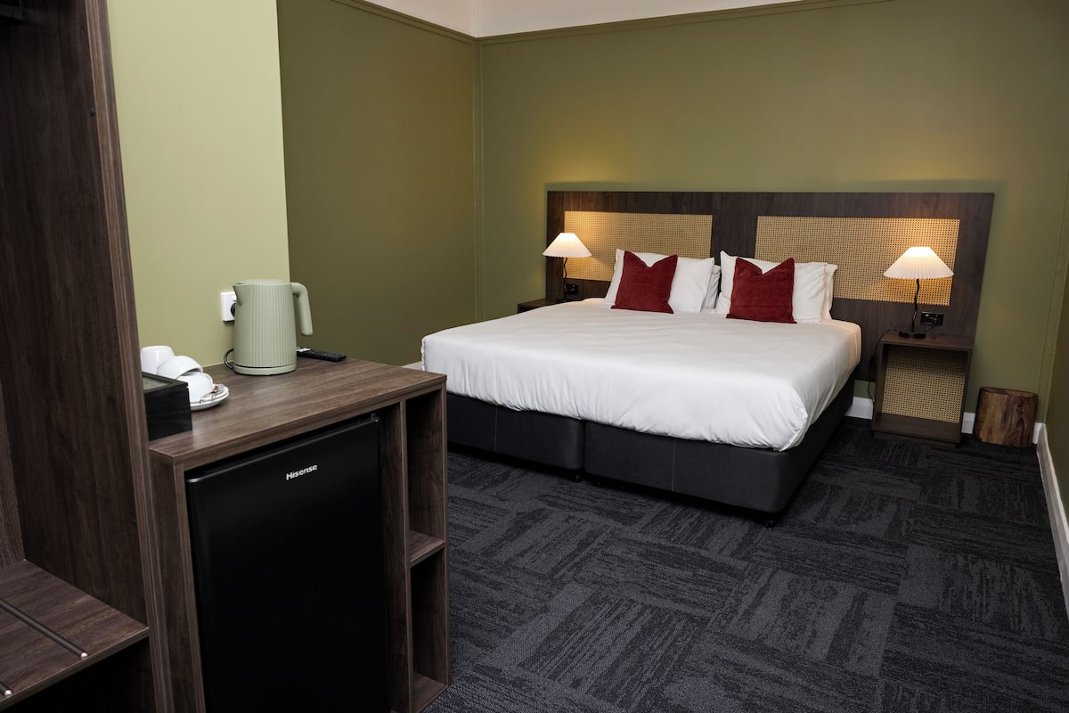 A hotel room with a bed, bedside lamps, a desk with a coffee maker, and a small refrigerator.