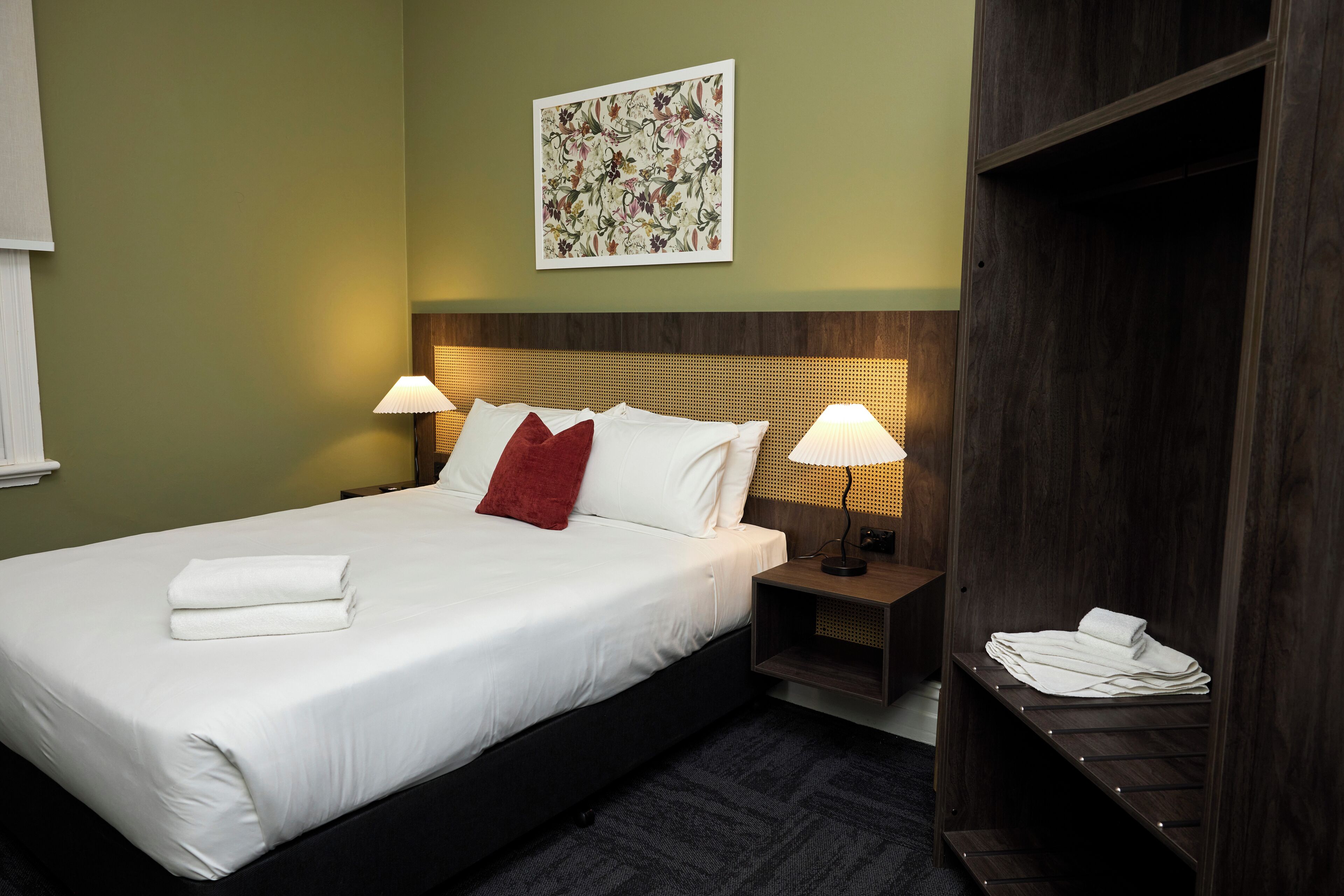 Un lit bien fait, avec du linge de lit blanc et un coussin rouge, deux lampes de chevet, une tête de lit en bois, une œuvre d’art florale encadrée au mur et une armoire en bois contenant des serviettes pliées.