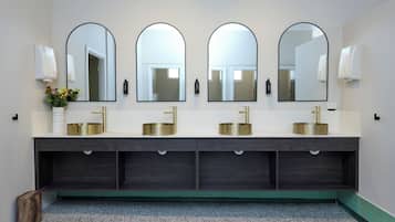 Un baño con tres lavamanos, cada uno con grifo dorado, y un espejo sobre ellos. Hay dos dispensadores de jabón empotrados en la pared a cada lado de los lavamanos.