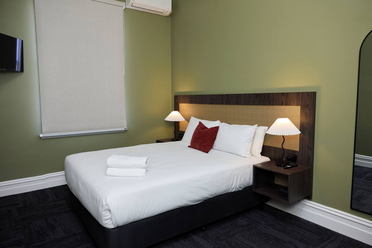 A neatly made bed with white linens and a red pillow, a bedside lamp, and a wall-mounted TV.