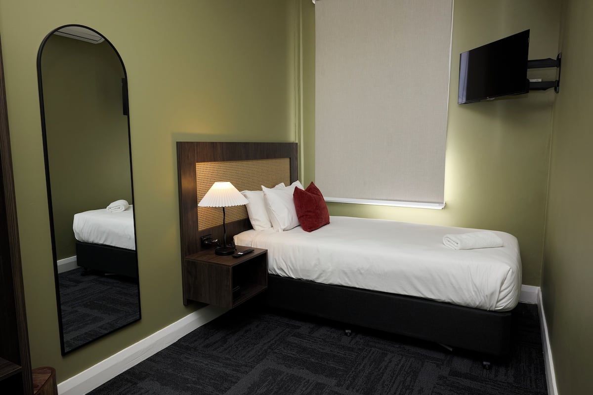 A hotel room with a single bed, a nightstand with a lamp, a TV mounted on the wall, and a large mirror.