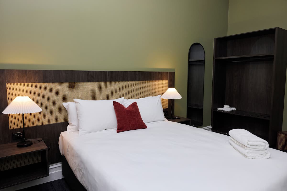 A hotel room with a neatly made bed, two bedside lamps, a red accent pillow, and a stack of towels.