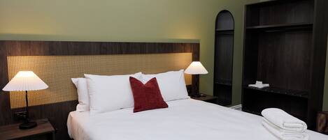 Habitación de hotel con una cama bien hecha, dos lámparas de noche, un cojín rojo decorativo y una pila de toallas.
