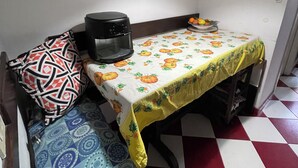 A compact room with a checkered floor, a table with a pumpkin-patterned tablecloth, a black appliance on the table, a patterned cushion on a chair, and a hanging light fixture.