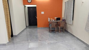 A hallway with a marble floor, a black door, and an orange wall. There is a table with chairs and a clock on the wall. - Sarvik Residency (Tiruchirappalli)