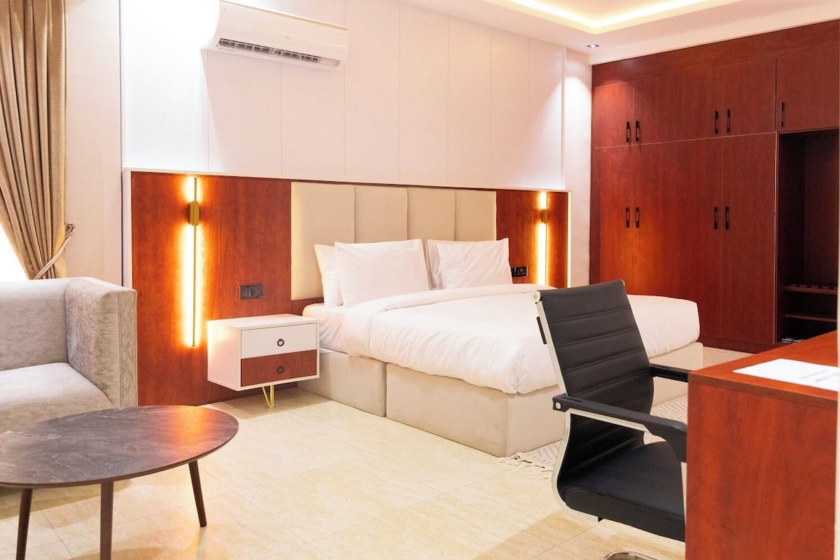 A modern hotel room with a bed, bedside tables, a desk, a chair, and a wardrobe.