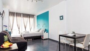 A modern hotel room with a bed, a dining table with chairs, a small table with fruit, a TV, and a window with curtains. - Villa Sagrada (Barcelona)