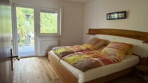 Un dormitorio con cama de madera, ventana con vistas a zonas verdes, radiador y puerta que da a un área exterior.