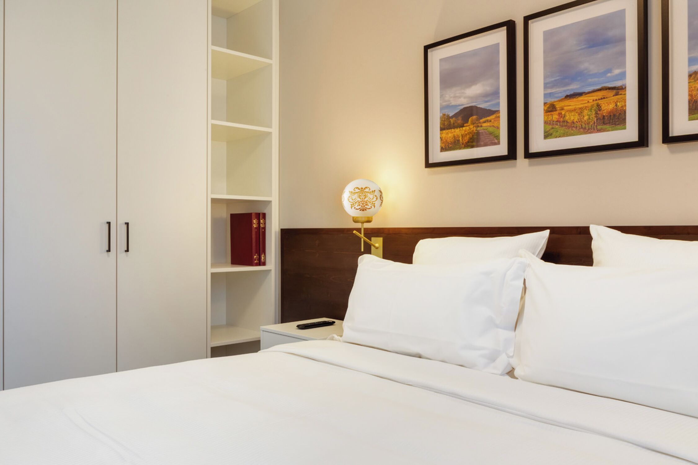 A hotel room with a bed, white bedding, a nightstand with a lamp, a wooden headboard, a bookshelf with books, and framed pictures on the wall.