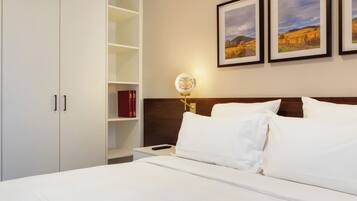 A hotel room with a bed, white bedding, a nightstand with a lamp, a wooden headboard, a bookshelf with books, and framed pictures on the wall.