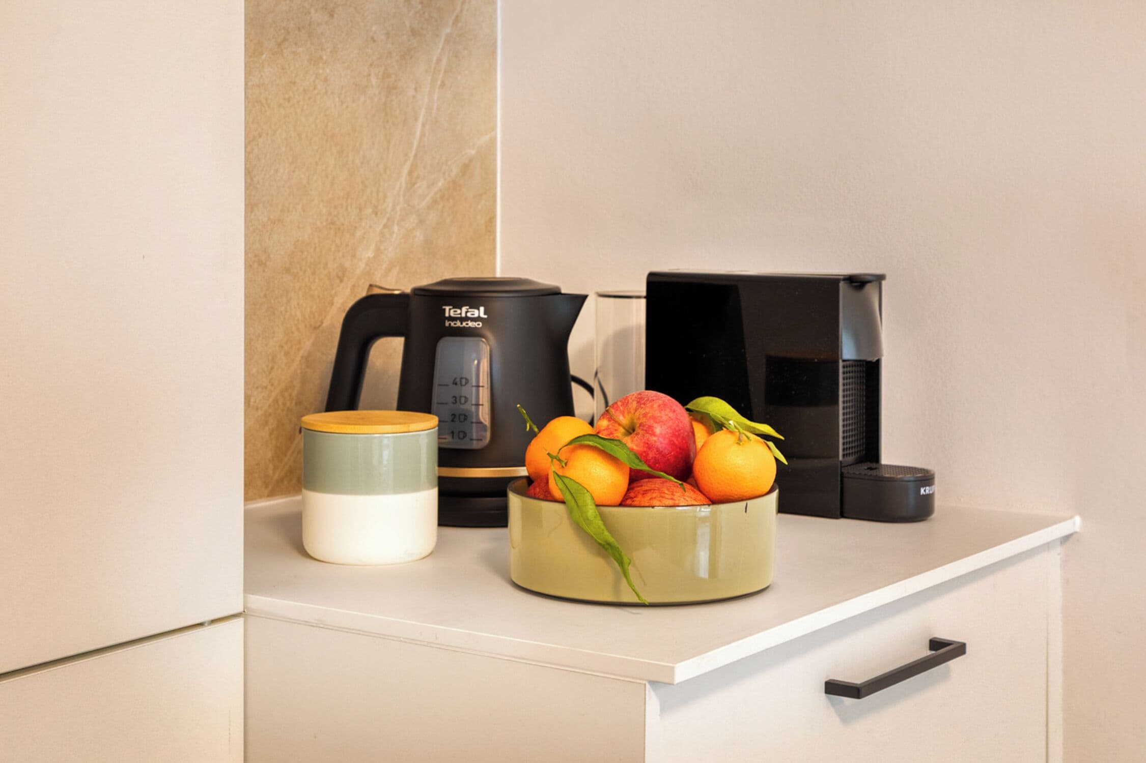 Una encimera de cocina con una cafetera, un cuenco de fruta y un recipiente.