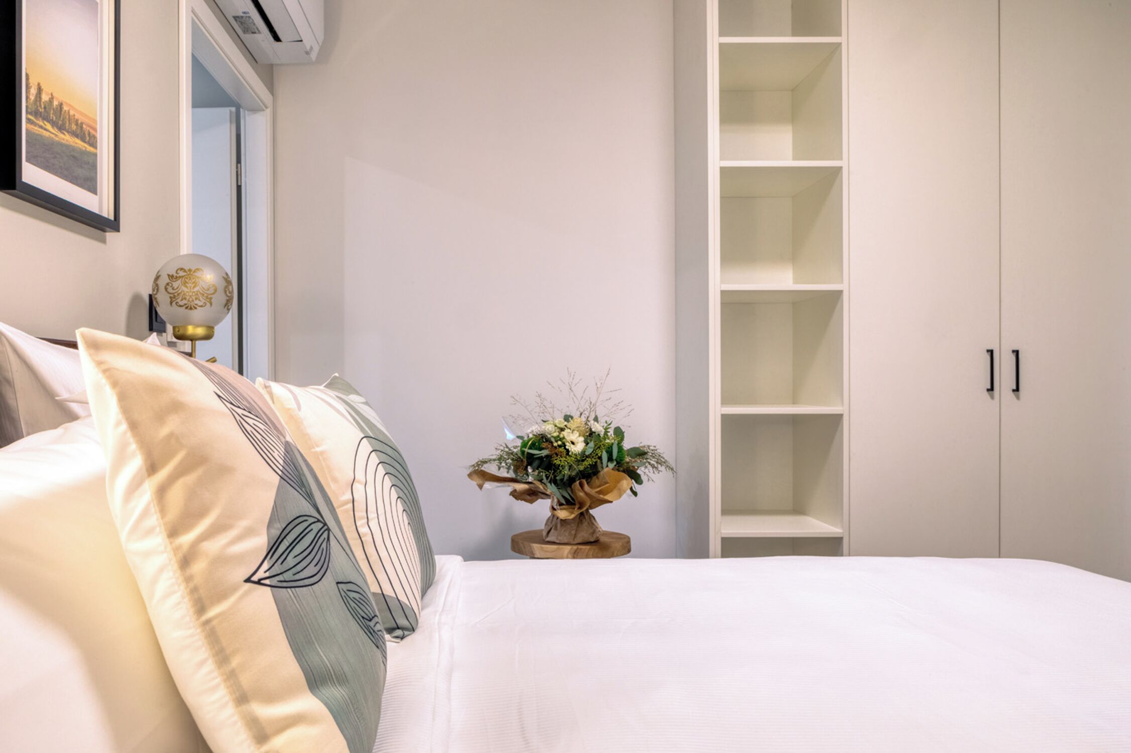 Un dormitorio con cama, mesita de noche con lámpara, aire acondicionado instalado en la pared y armario blanco con puertas abiertas.