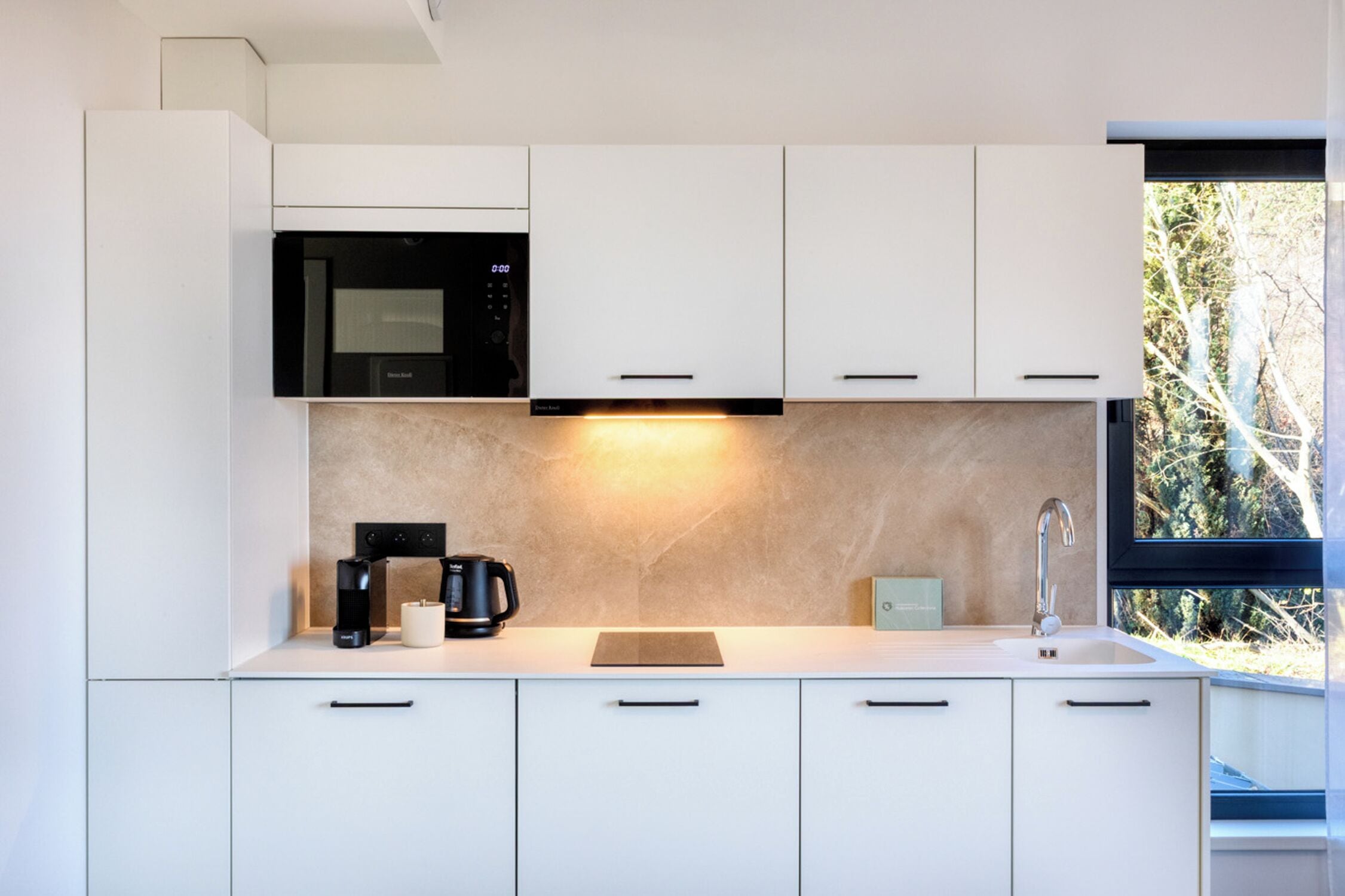 A modern kitchen with white cabinets, a built-in microwave, a coffee maker, and a window with a view of trees.