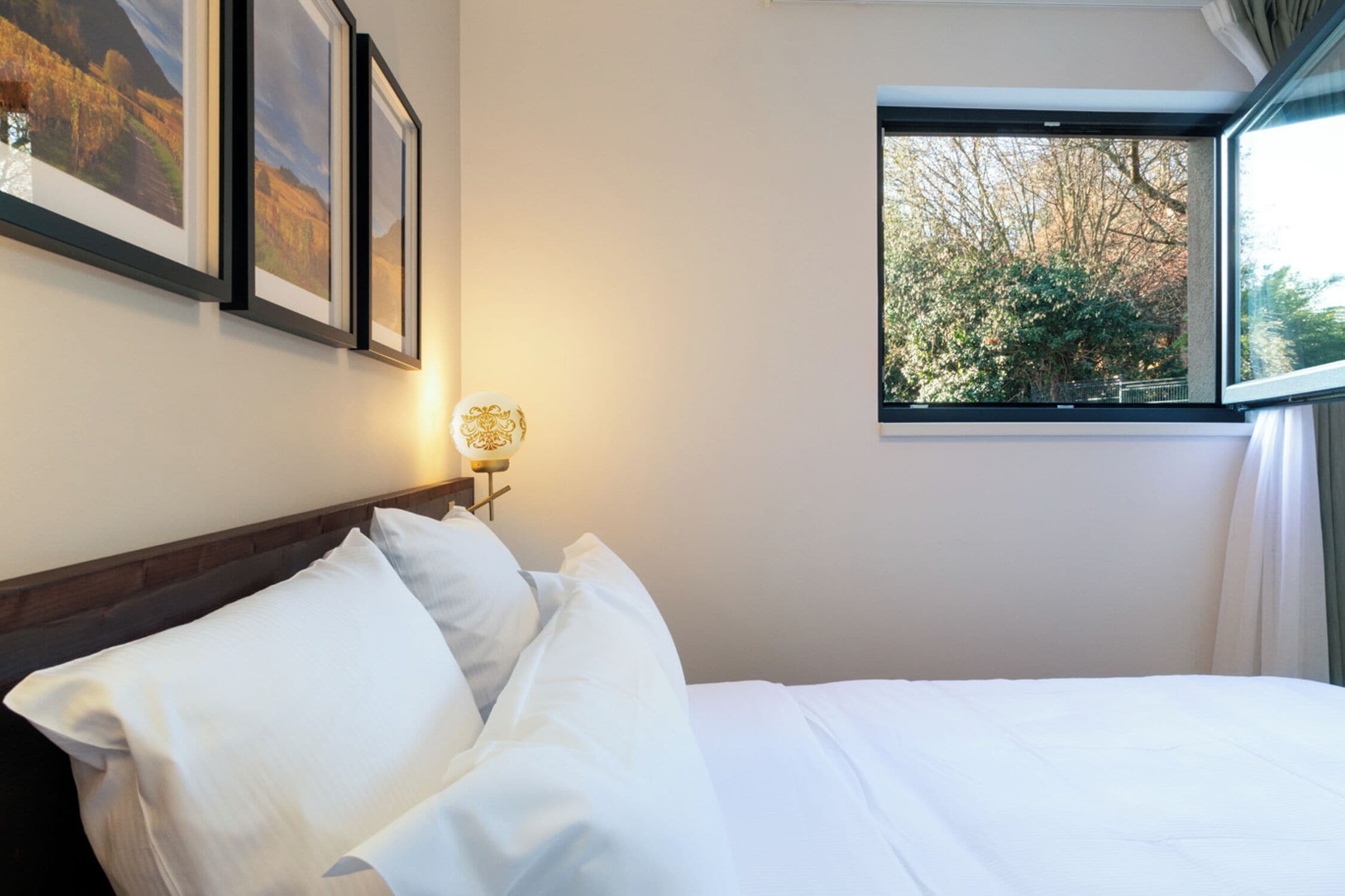 Ein Hotelzimmer mit einem ordentlich bezogenen Bett, gerahmten Bildern an der Wand, einer Nachttischlampe und einem Fenster mit Blick auf Bäume.