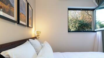 Ein Hotelzimmer mit einem ordentlich bezogenen Bett, gerahmten Bildern an der Wand, einer Nachttischlampe und einem Fenster mit Blick auf Bäume.