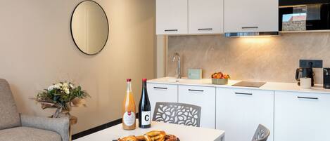 A modern kitchen with white cabinets, a built-in oven, and a sink. There is a dining table with pretzels and a bottle of wine.