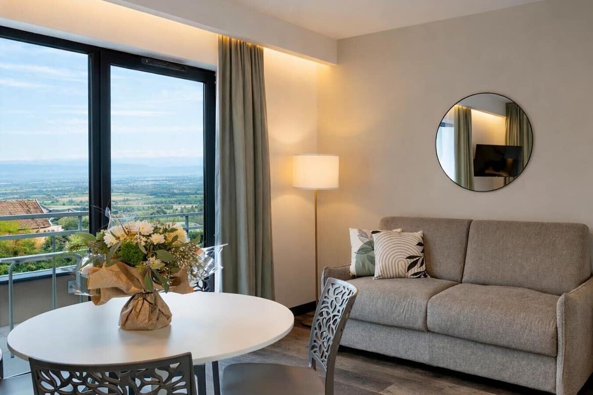 A modern hotel room with a sofa, a round mirror, a dining table with chairs, and a view of a cityscape through large windows.
