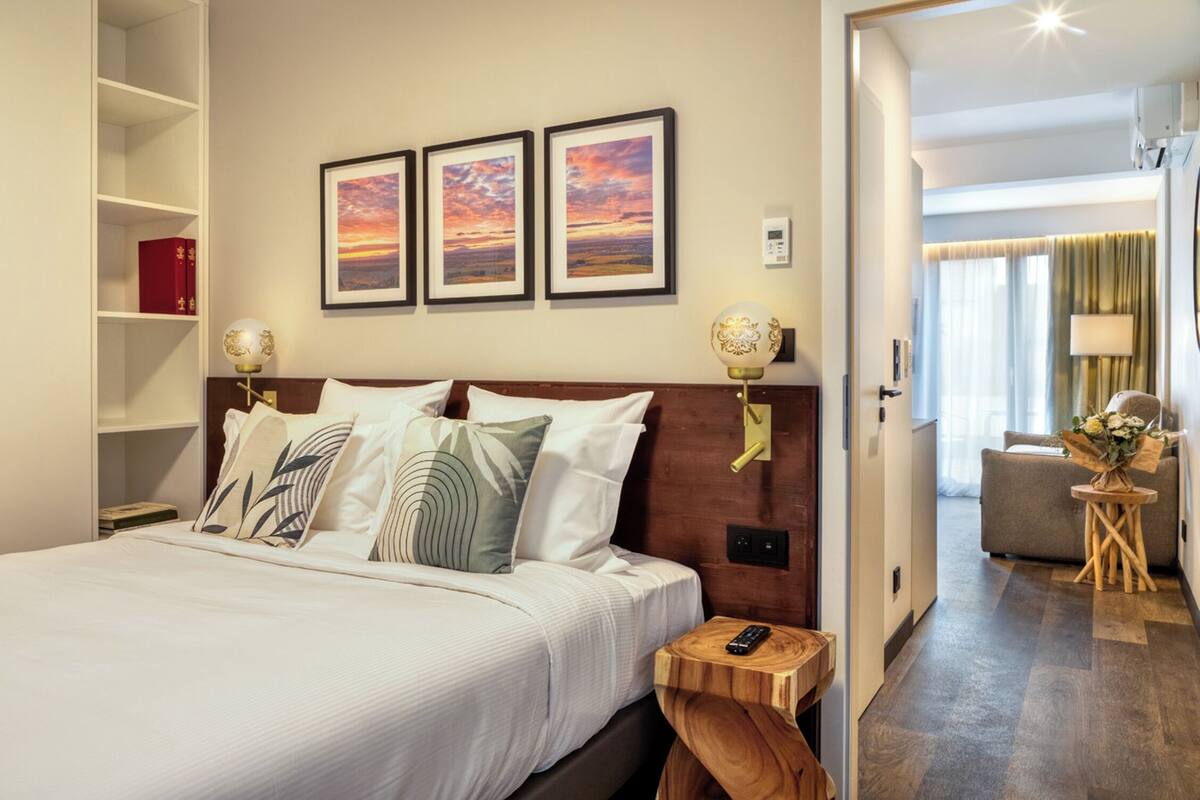 A hotel room with a large bed, a wooden headboard, and a view of the hallway. There are three framed pictures on the wall, a bedside table with a lamp, and a small wooden stool.