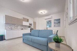 A modern living room with a sofa, a small round table with a plant, and a kitchen in the background.