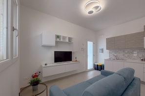A modern living room with a sofa, a TV on a white cabinet, a small table with a plant, and a kitchen area with cabinets and a sink.