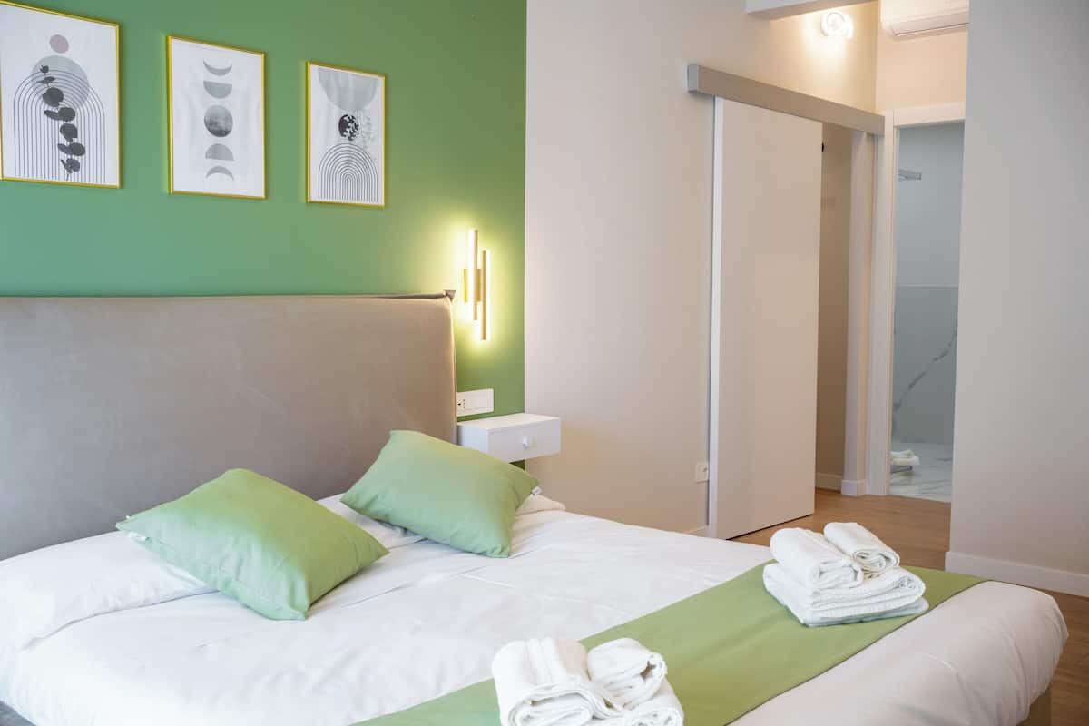 A neatly made bed with green pillows, a green throw blanket, and rolled towels on the side. There are framed artworks on the wall above the bed.