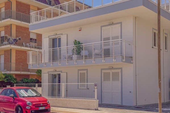 A multi-story residential building with balconies, a red car parked in front, and a clear sky.