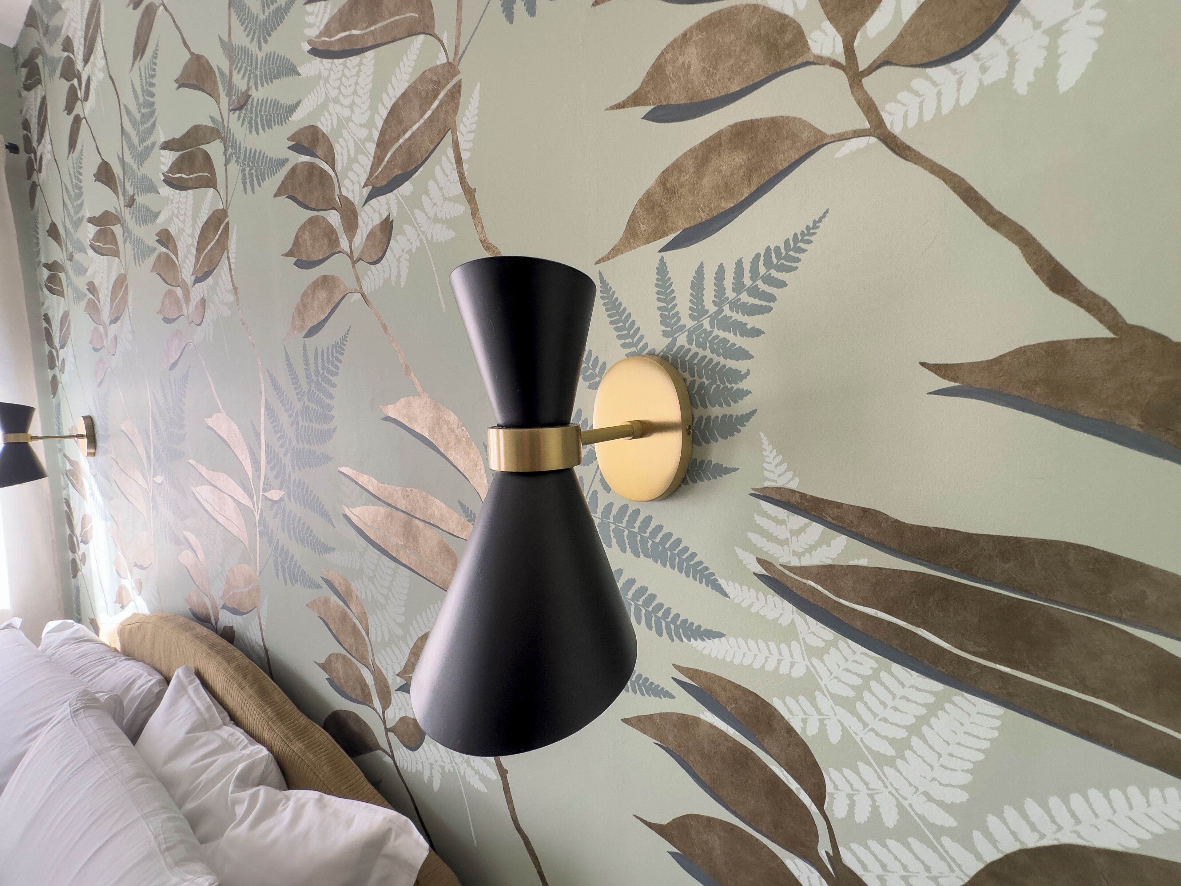 A room with a bed, a wall-mounted lamp, and a floral wallpaper design.