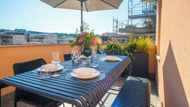 An outdoor dining area with a table set for two, an umbrella, and potted plants. - Terrace Apartment Monte del Gallo (Rome)