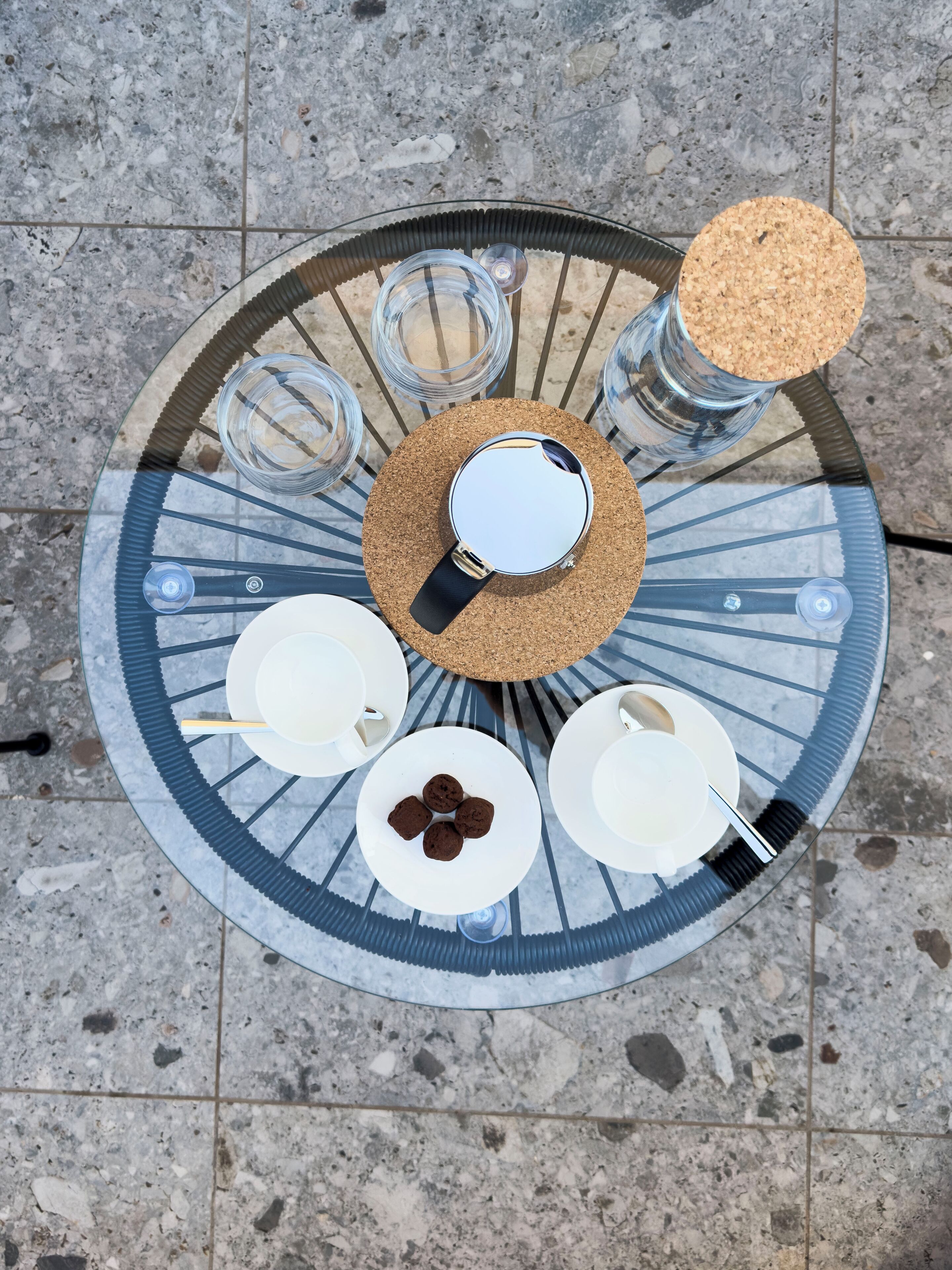 Een ronde glazen tafel met een kurken onderzetter, vier witte borden en vier glazen.