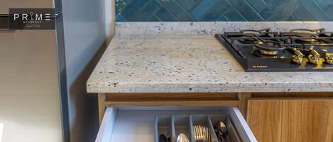 A kitchen with a stainless steel refrigerator, a granite countertop, and a drawer organizer with utensils.