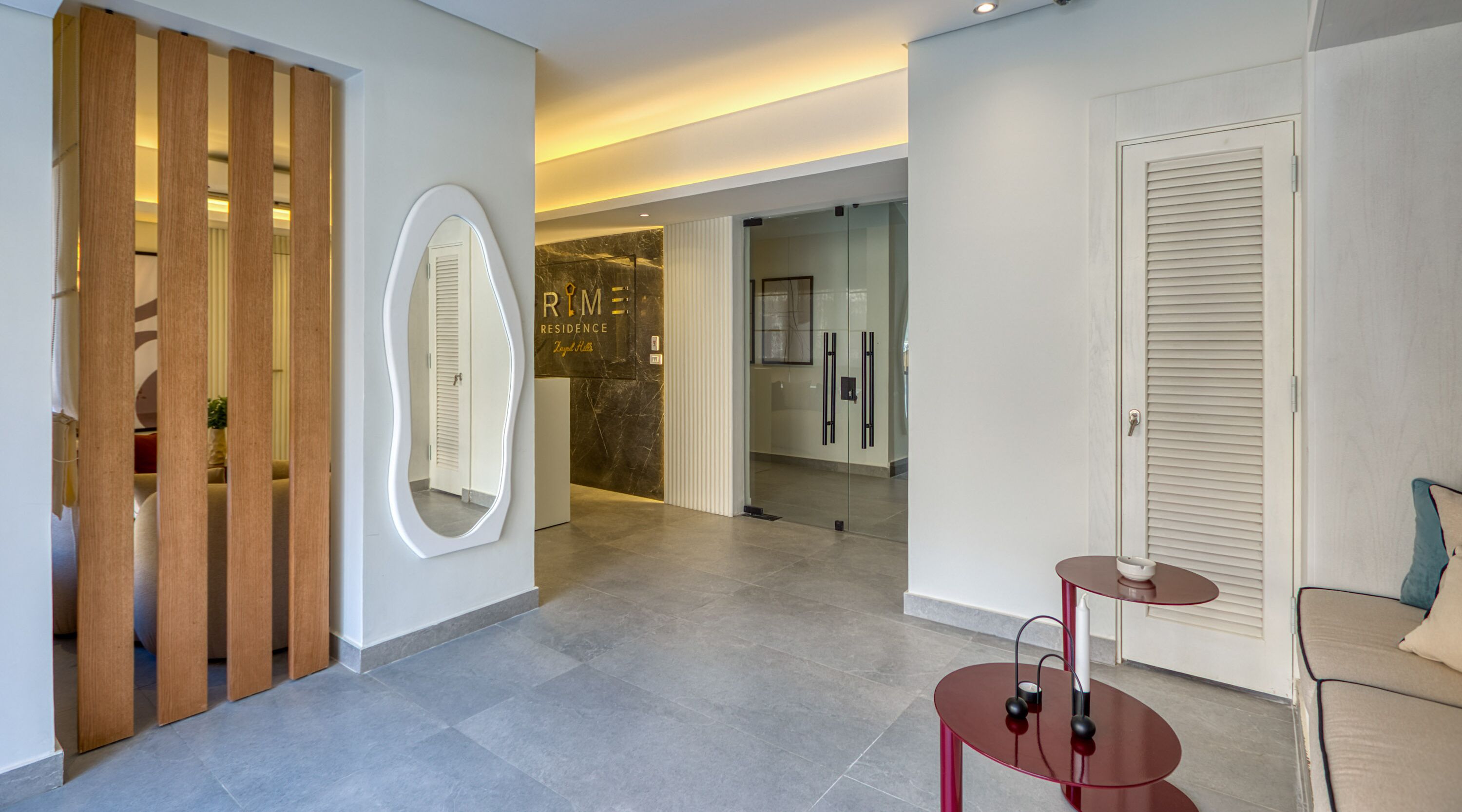 Un moderno vestíbulo de hotel con un separador de madera, un espejo grande, una puerta de vidrio que da a un baño y una zona de estar con una mesa roja y un sofá.