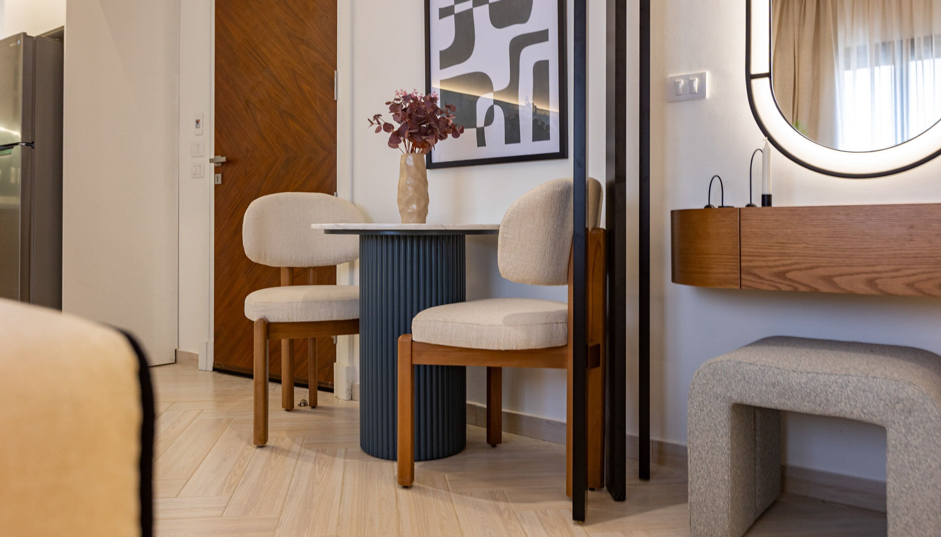 Une chambre d’hôtel moderne avec une porte en bois, une table d’appoint bleue ornée d’un vase, deux chaises beiges, un meuble en bois et un miroir rond.