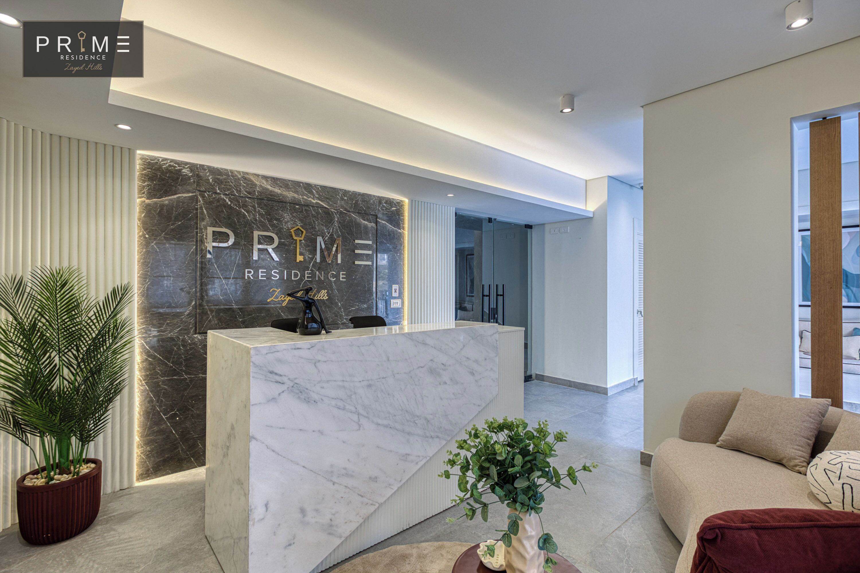 Um balcão de recepção moderno com tampo de mármore e uma placa que diz "PRIME RESIDENCE". Há uma planta em vaso à esquerda e um sofá com um cobertor por perto.