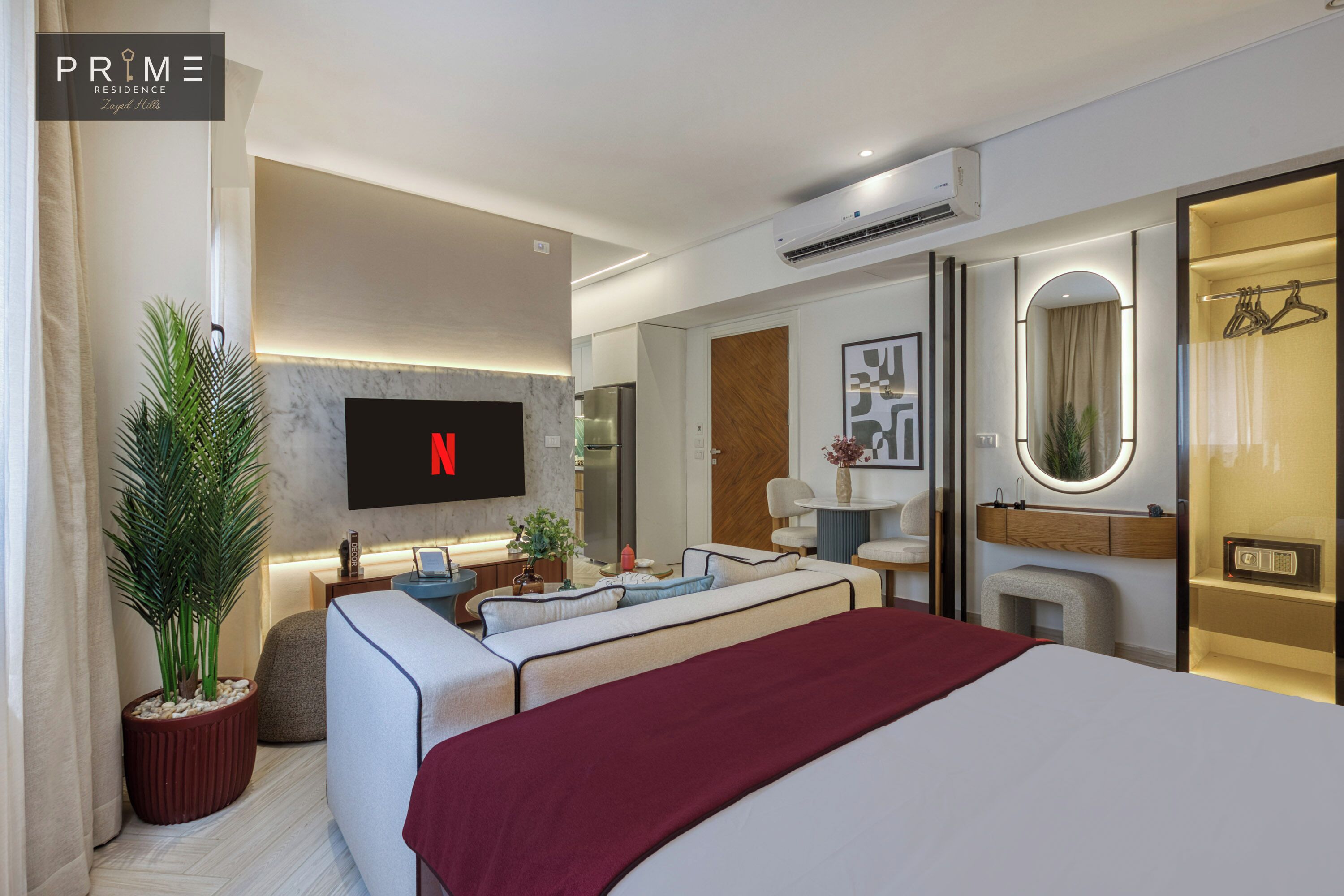 A modern hotel room with a large bed, a TV mounted on the wall, a seating area with a sofa and armchairs, a small table, and a mirror with a wooden frame.