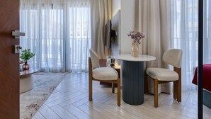 A modern living room with a round table, two chairs, a TV mounted on the wall, and sheer curtains. - Prime Residence Zayed Hills (Sheikh Zayed City)