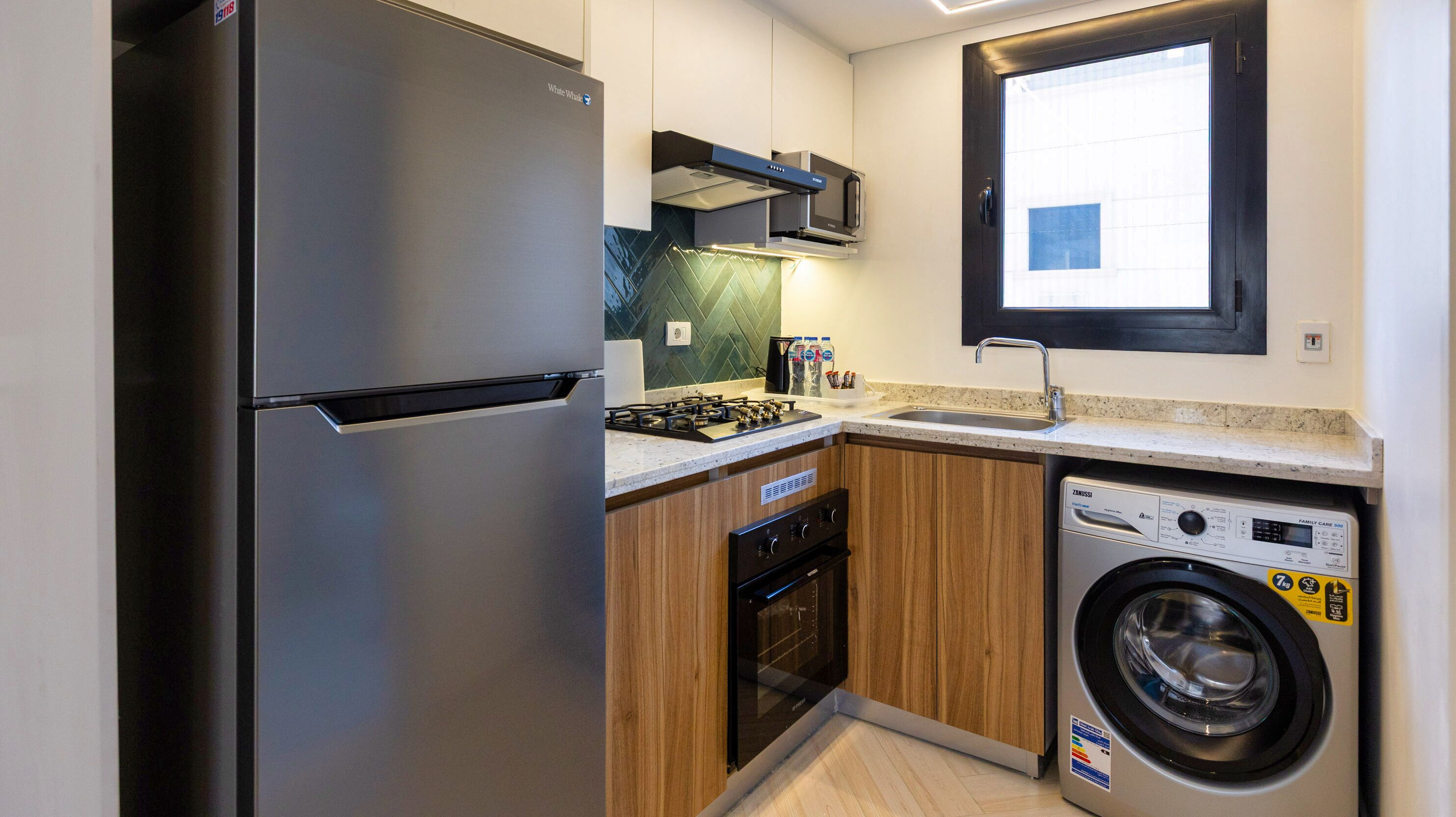 A compact kitchen with a stainless steel refrigerator, a built-in oven, a washing machine, and a window above the sink.