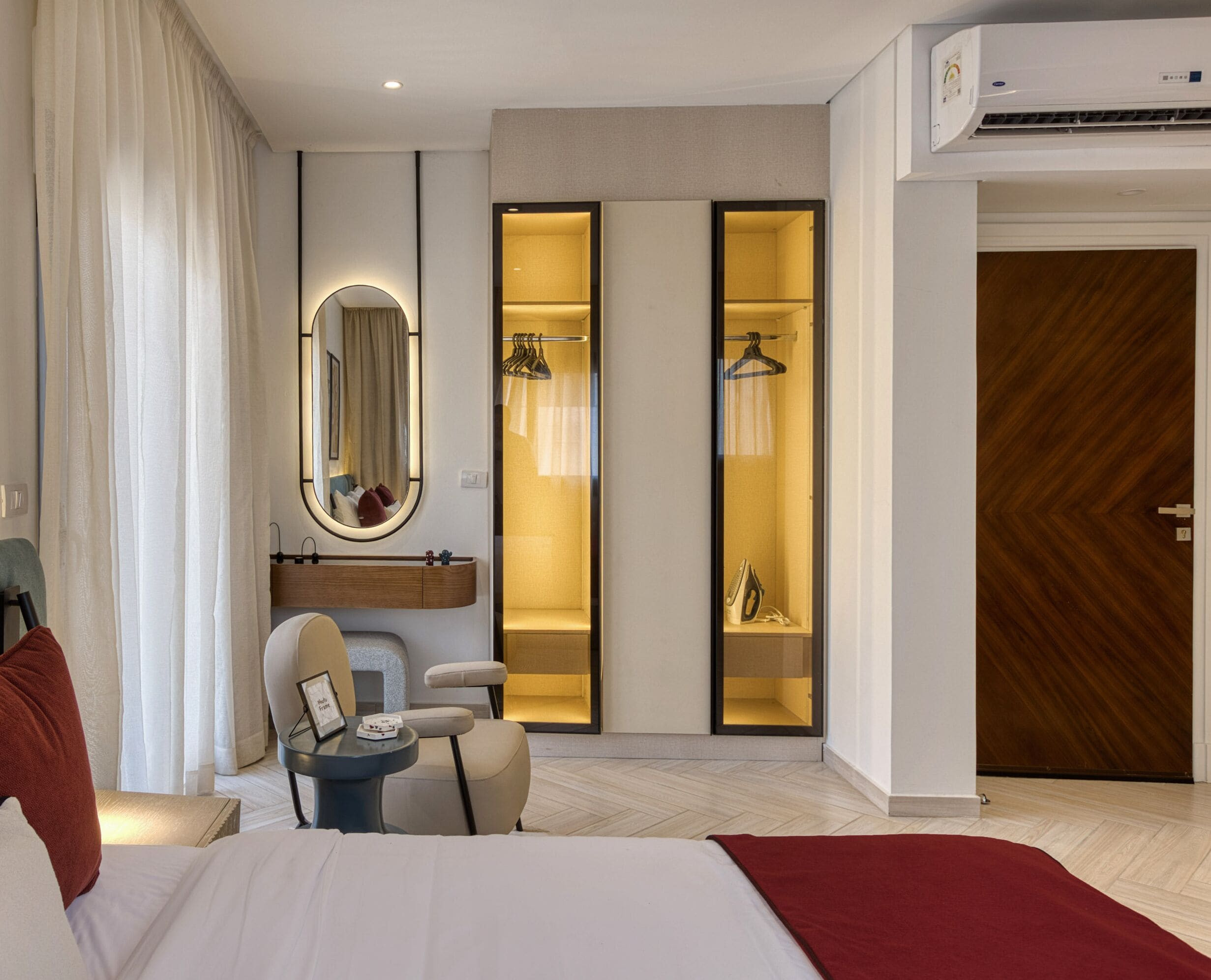 A hotel room with a bed, a desk, a chair, a mirror, a wardrobe, and an air conditioning unit.